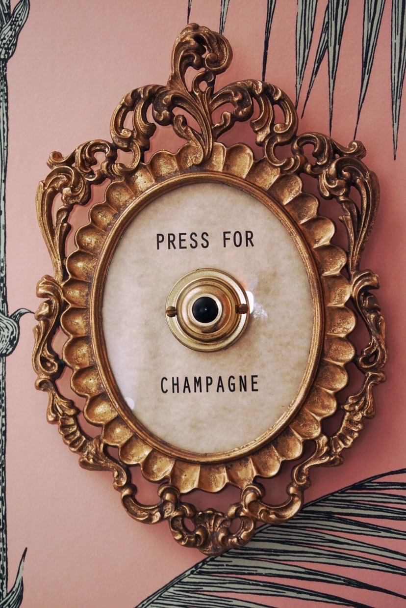 An ornate gold bell on pink wallpaper reading Press for Champagne